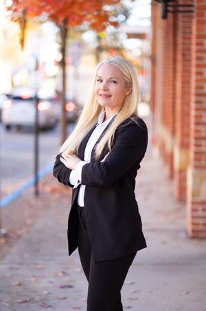 Professional woman in black business suit stands confidently on autumn sidewalk with orange fall foliage in background.