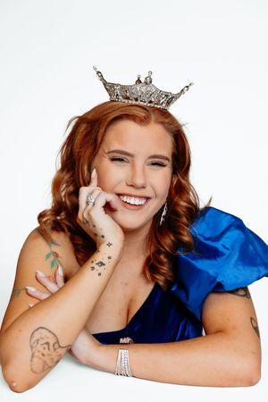 Smiling young woman with red hair wearing a silver crown and blue dress, showing tattoos on her arms.