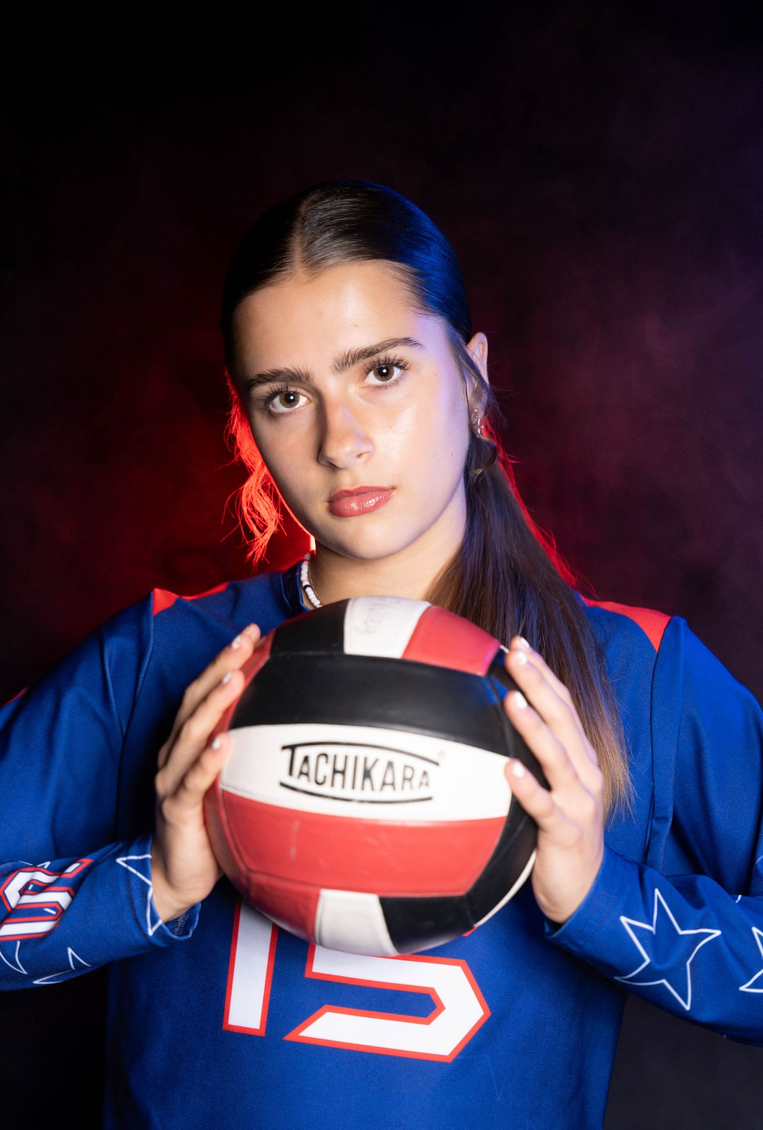 Volleyball player in blue jersey holds red and white volleyball against dark background.