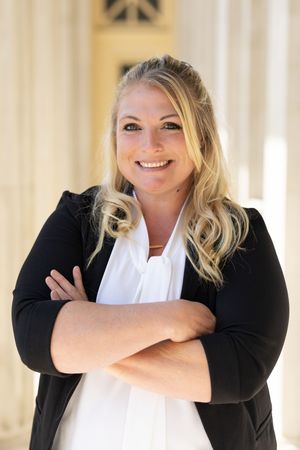 Professional woman in black blazer and white blouse smiling with arms crossed in front of elegant columns.