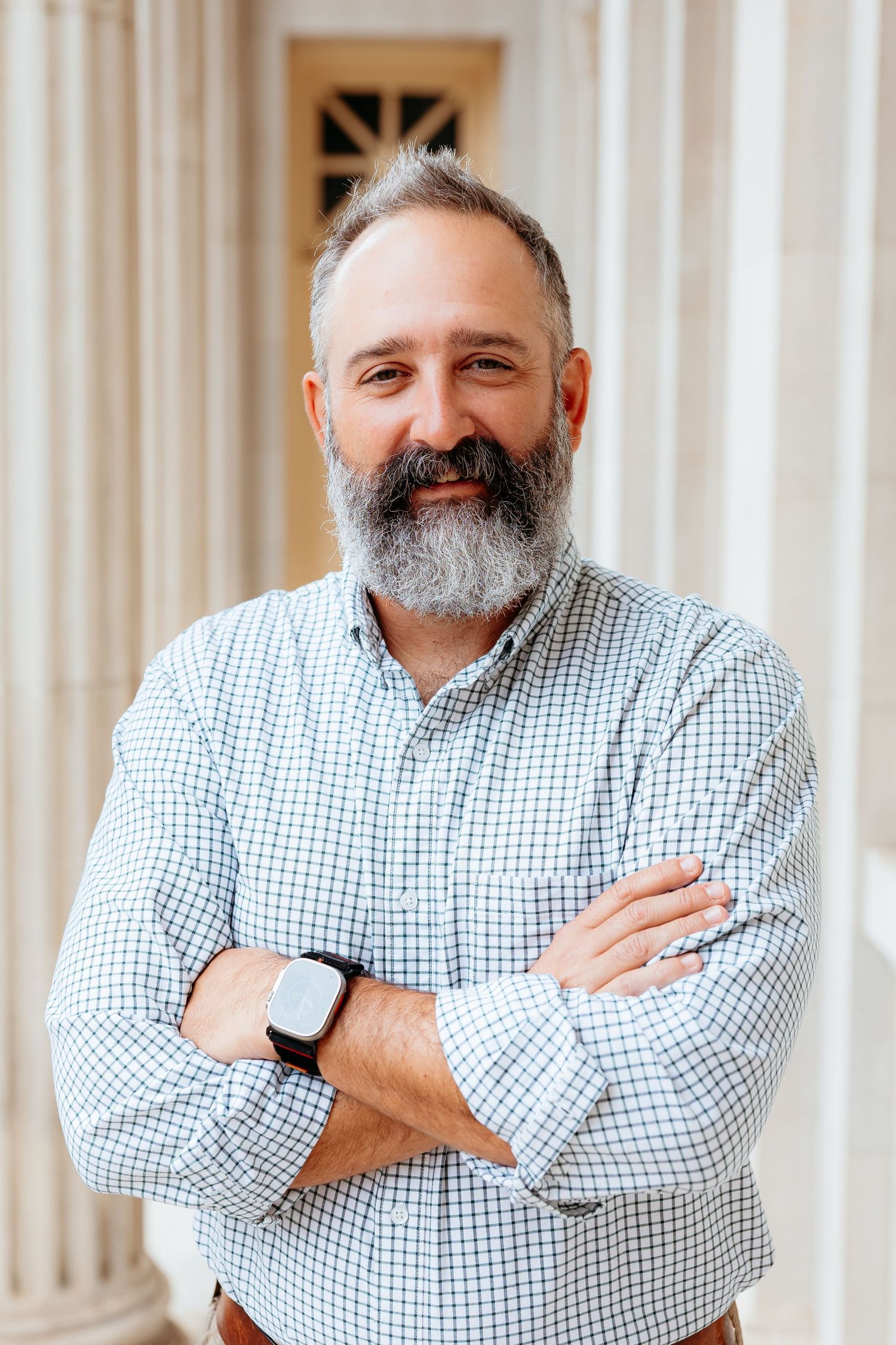 Smiling middle-aged man with gray beard, arms crossed, wearing a checkered shirt and smartwatch, standing near white columns.