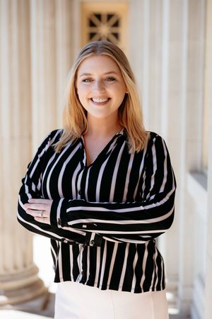Young blonde woman in striped black and white blouse stands confidently with arms crossed, smiling before classic columns.