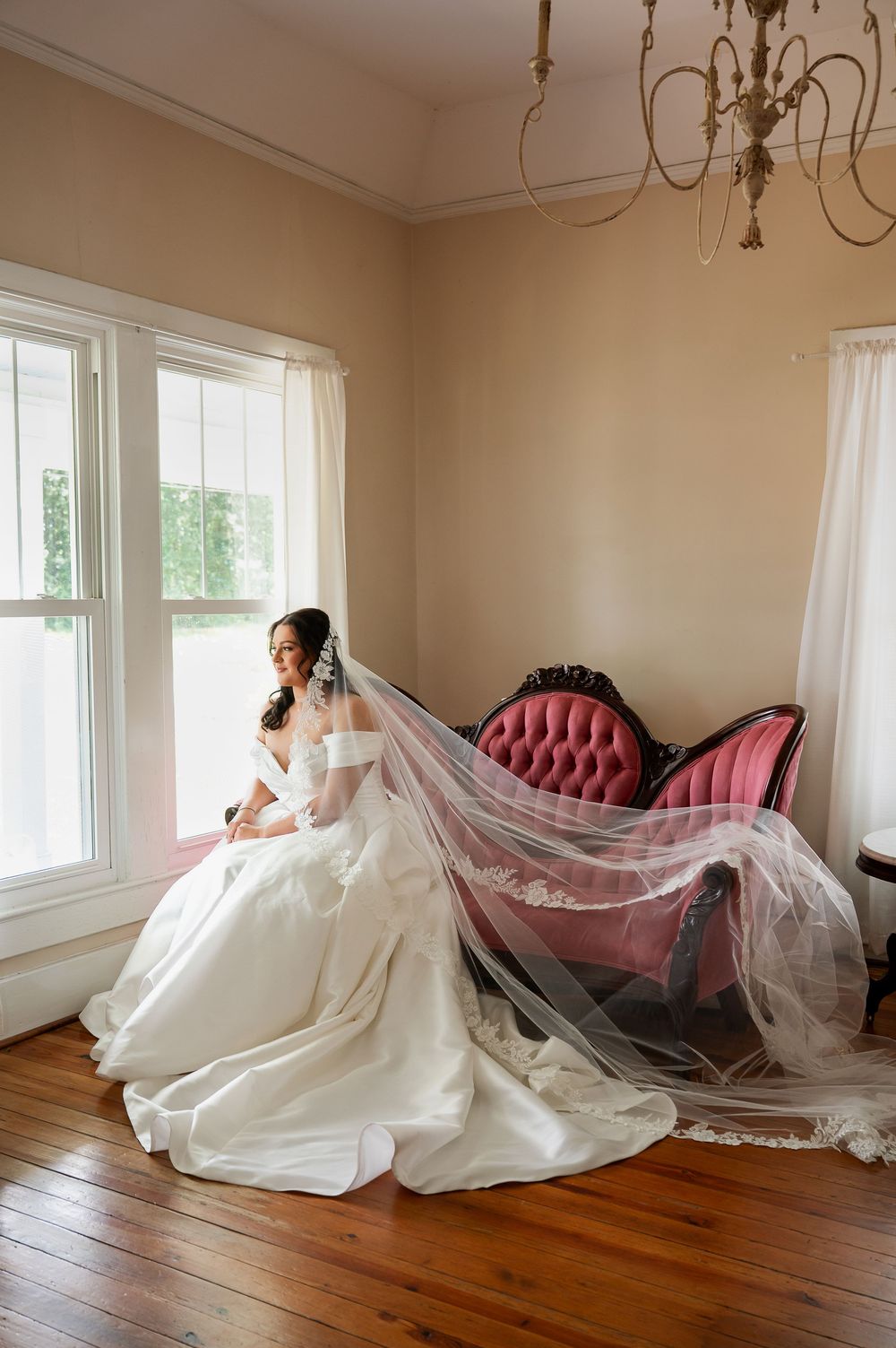 A wedding dress photoshoot featuring a pink chaise lounge and flowing veil in a sunlit room.
