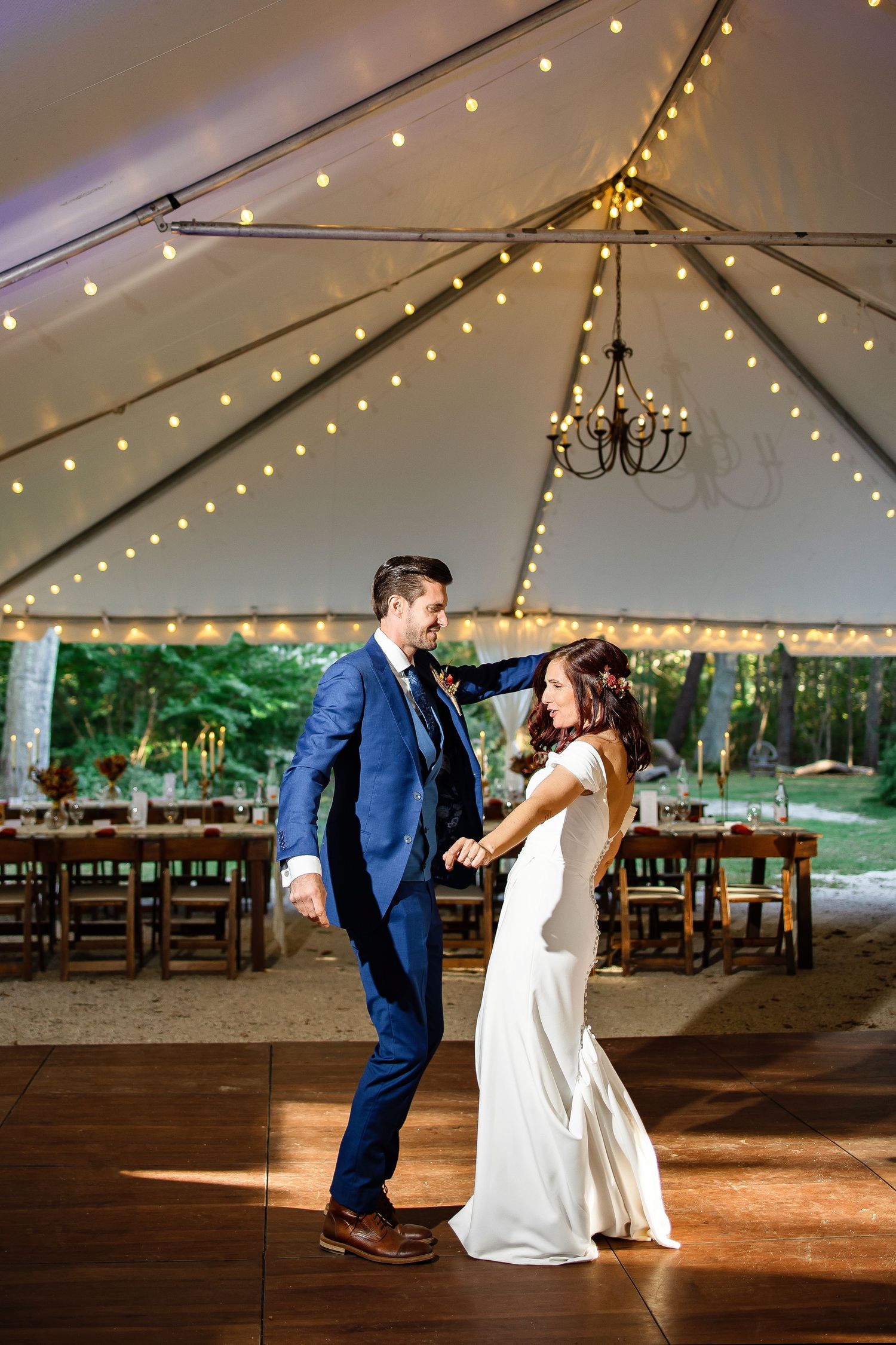 A couple shares their first dance under a tent with string lights and chandeliers at an outdoor wedding reception.