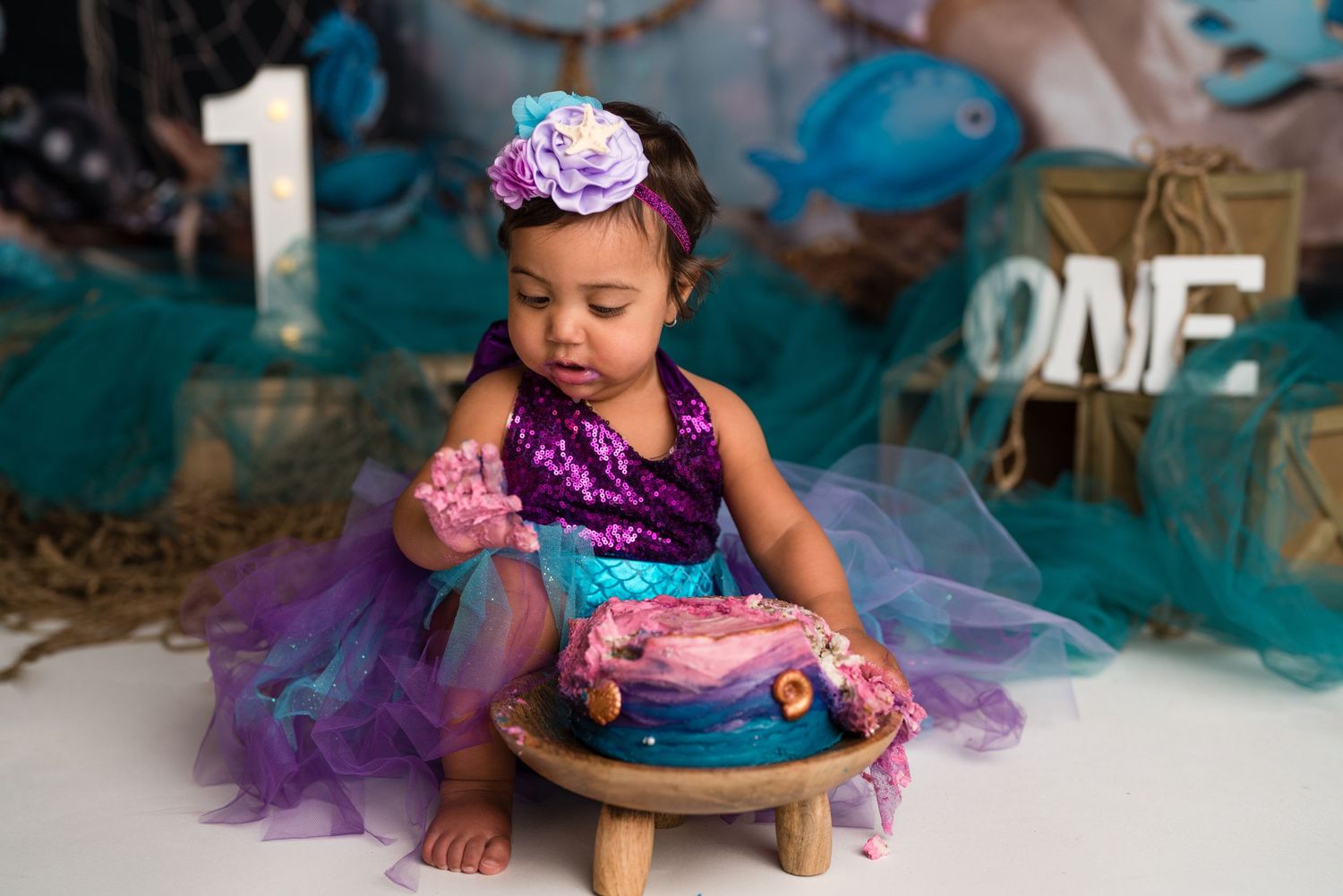 A cake smash photoshoot features purple and turquoise decorations with a small cake on a wooden stool.