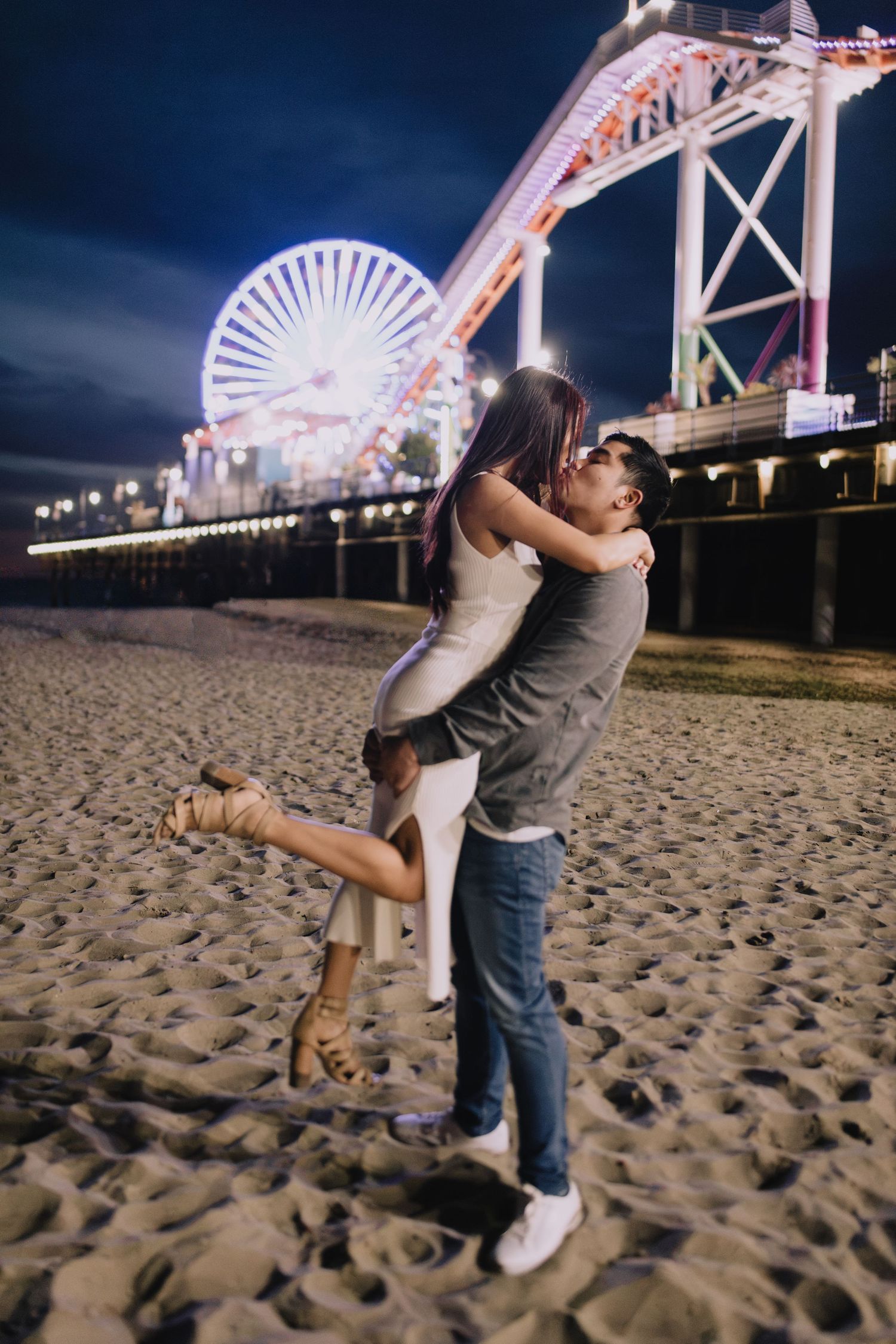 Man lifting woman in joyful embrace on Santa Monica beach at night with vibrant pier lights behind them.
