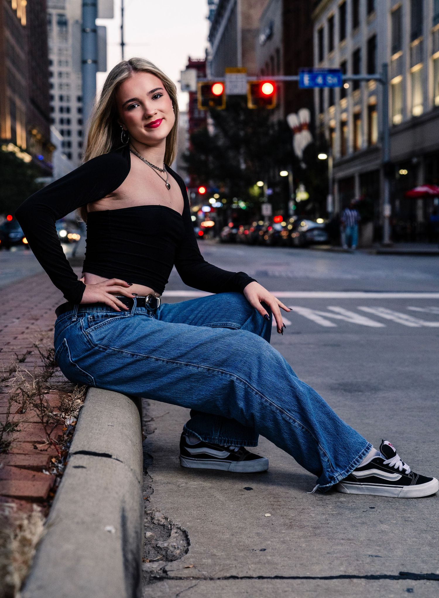 A person wearing jeans and black sneakers sits casually on a city curb at dusk with urban buildings in the background.