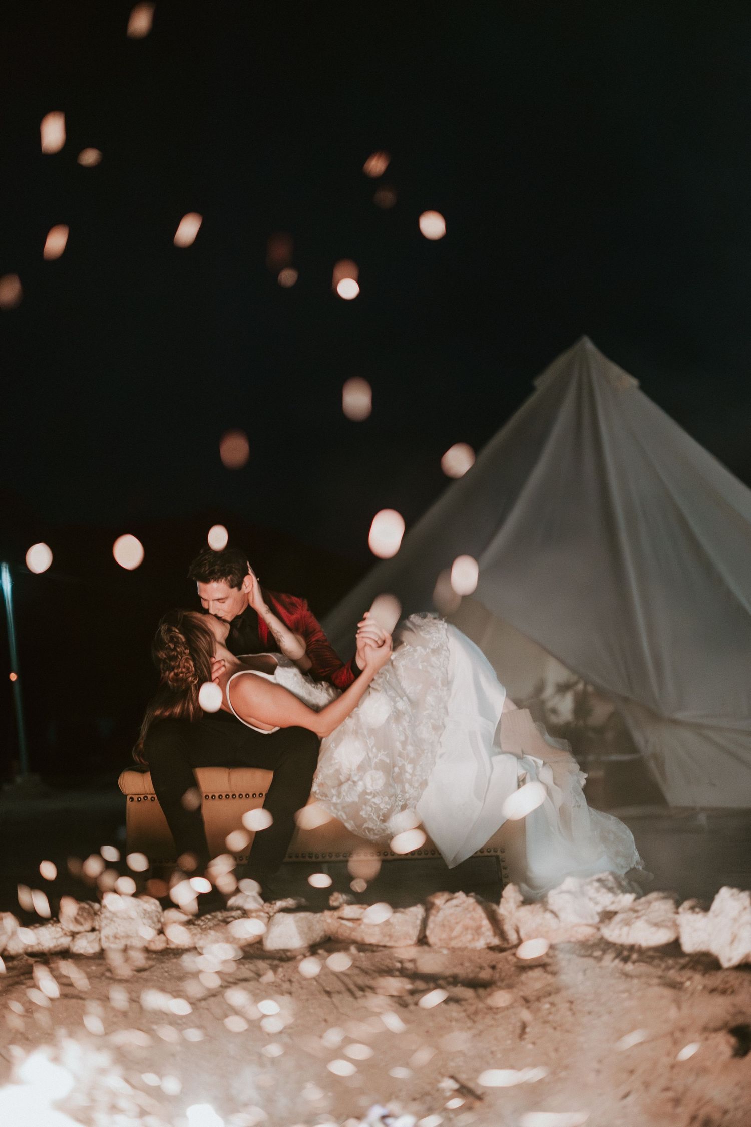 Couple shares romantic moment by glowing tent at night with floating lanterns illuminating the dark sky.