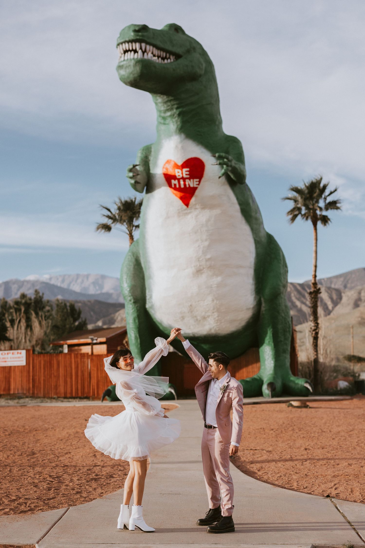 Couple in wedding attire dancing before giant green T-Rex dinosaur statue in Palm Springs.