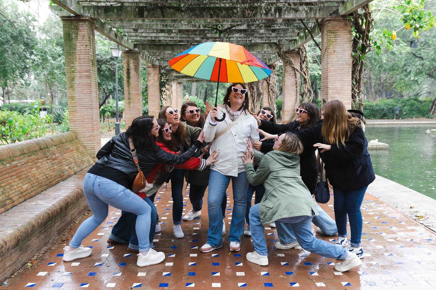 A group of people huddle together under a colorful umbrella during a rainy day at a park.
