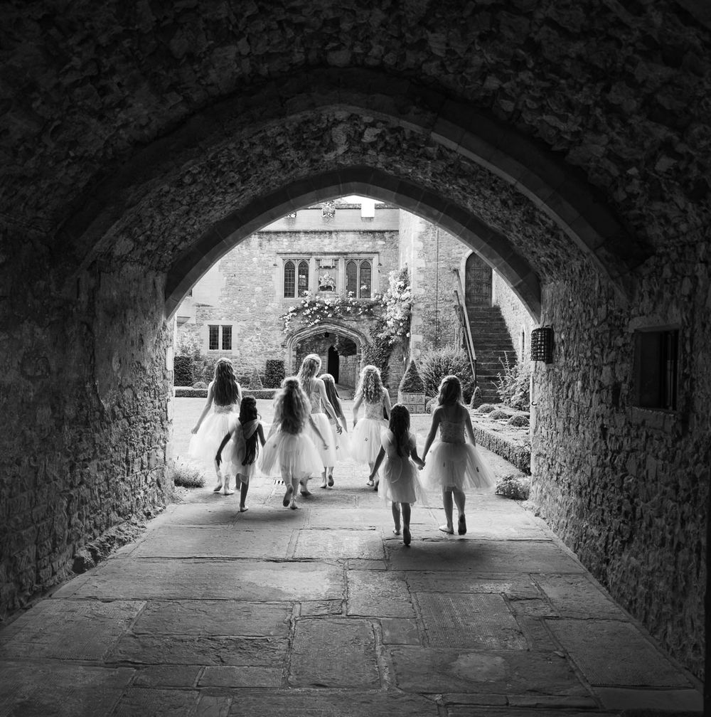 Flower girls dressed as ballet fairies walking through stone tunnel archway at Allington Castle, in black and white.