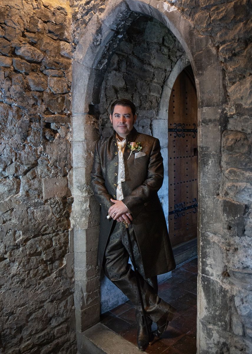 Groom dressed as prince in an ancient stone archway corridor with rough textured walls and dramatic lighting. Allington Castle.