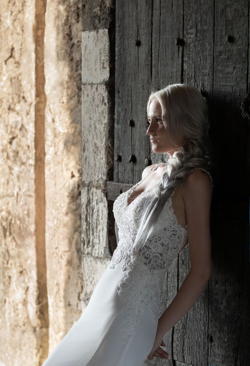 A bride in a white lace wedding dress leans against a rustic wooden wall in dramatic lighting | Allington Castle in Kent