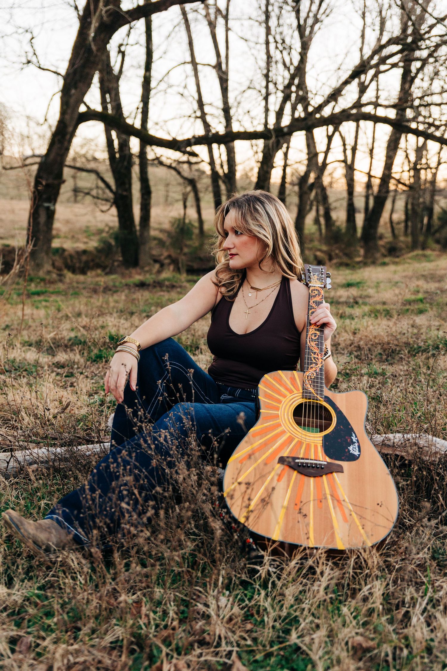 A guitarist sits in a rustic outdoor setting at sunset with an acoustic guitar among dried grass and bare trees.