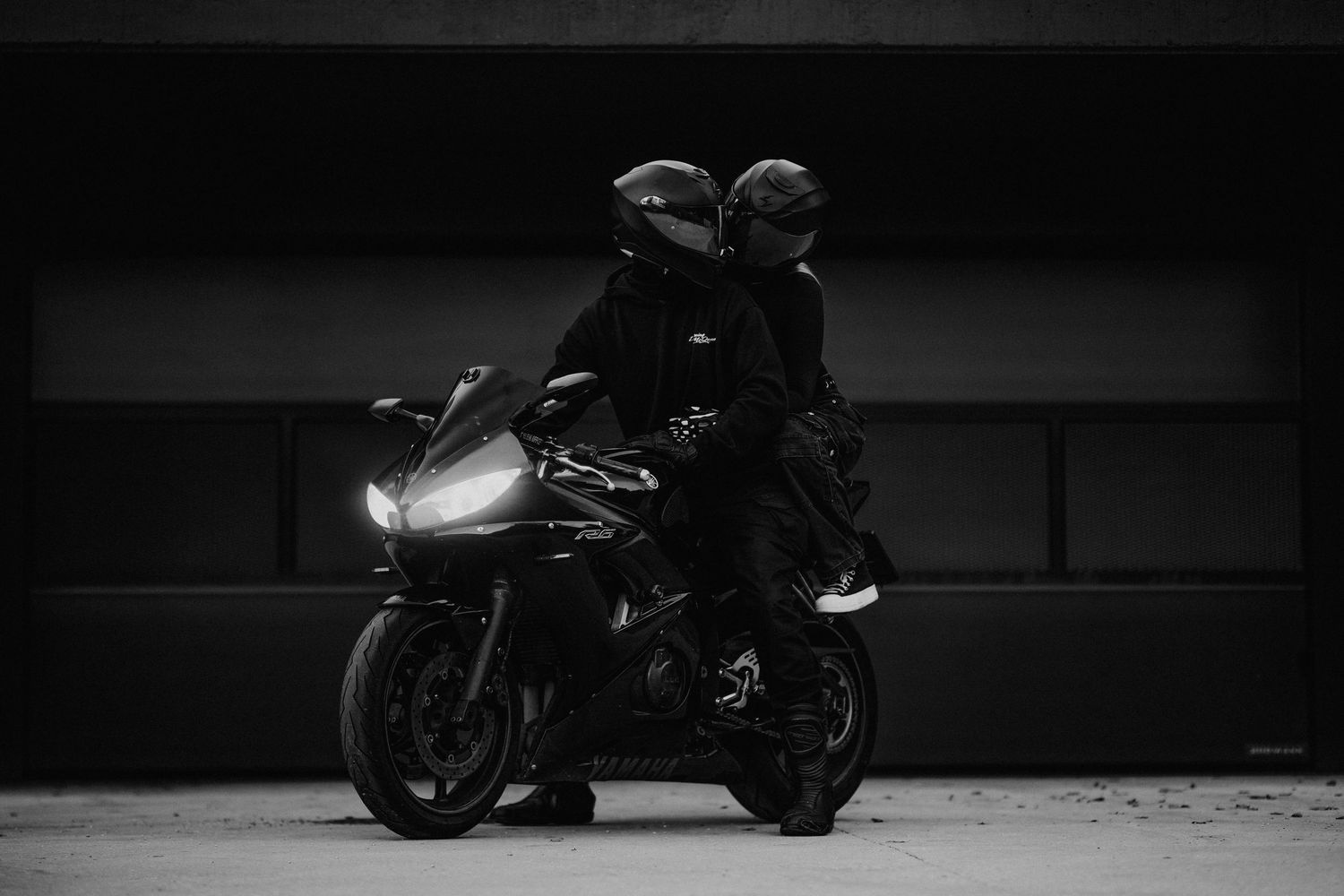 Umelecký široký záber na pár sediaci na športovej motorke v mestskom prostredí, dramatická čiernobiela kompozícia.