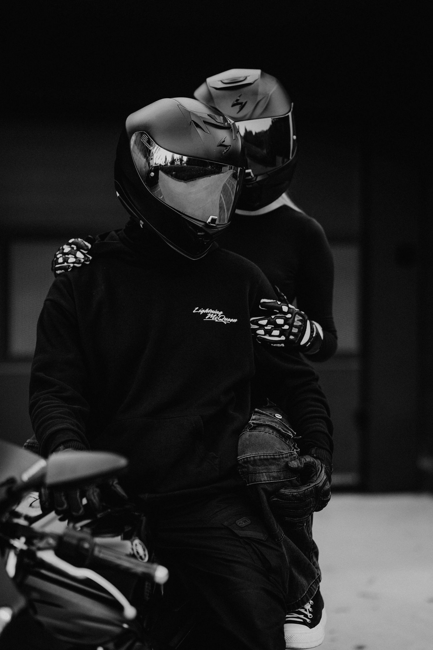 Detailný čiernobiely portrét páru v motorkárskom oblečení s prilbami, zachytenie spoločnej vášne a štýlu.