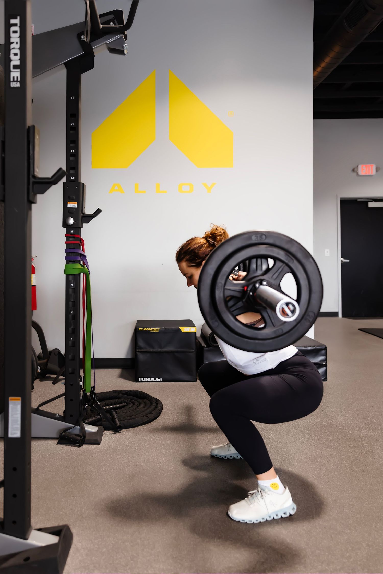 Person performing a squat exercise with a weight plate at a gym with yellow logo on wall.