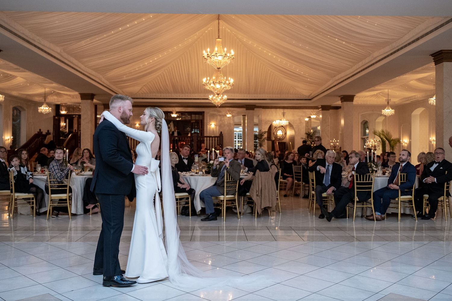 A wedding couple shares their first dance on a white dance floor under elegant chandeliers in a reception hall.
