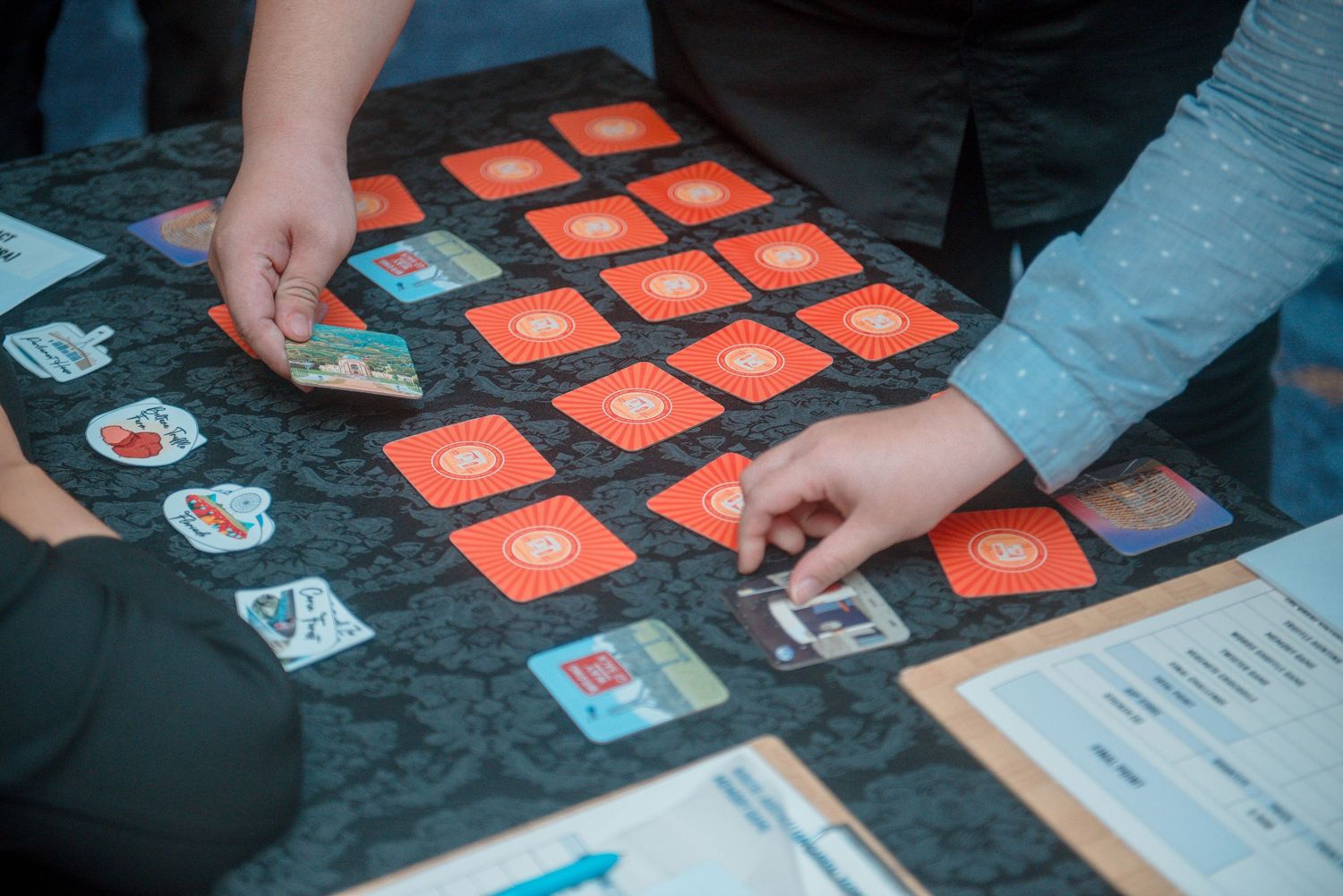 Multiple hands playing a card game on a dark surface with orange and white cards spread out.