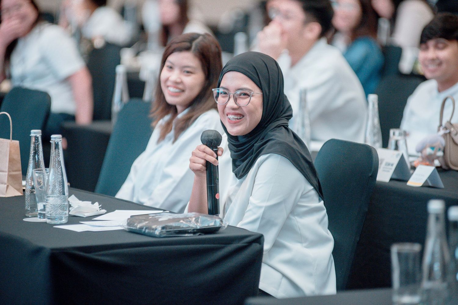 Attendees sit at tables during a professional conference with water bottles and microphones visible.