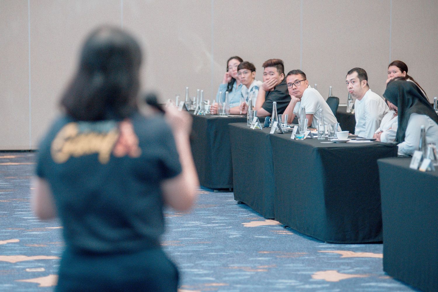 Business professionals sit at long tables during a conference or meeting in a modern office setting.