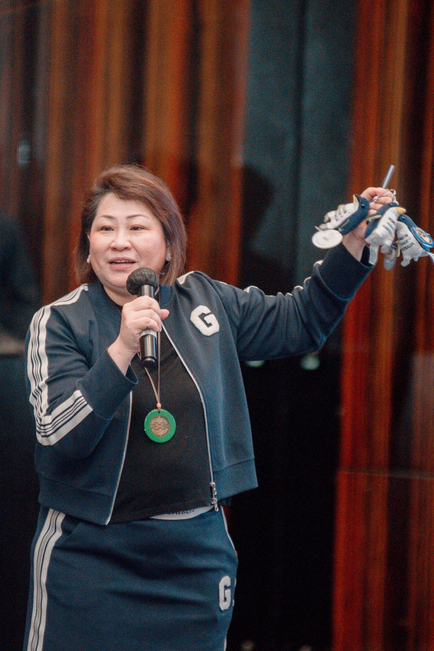 A person in a black tracksuit performs at a microphone stand with animated gestures and expressions.