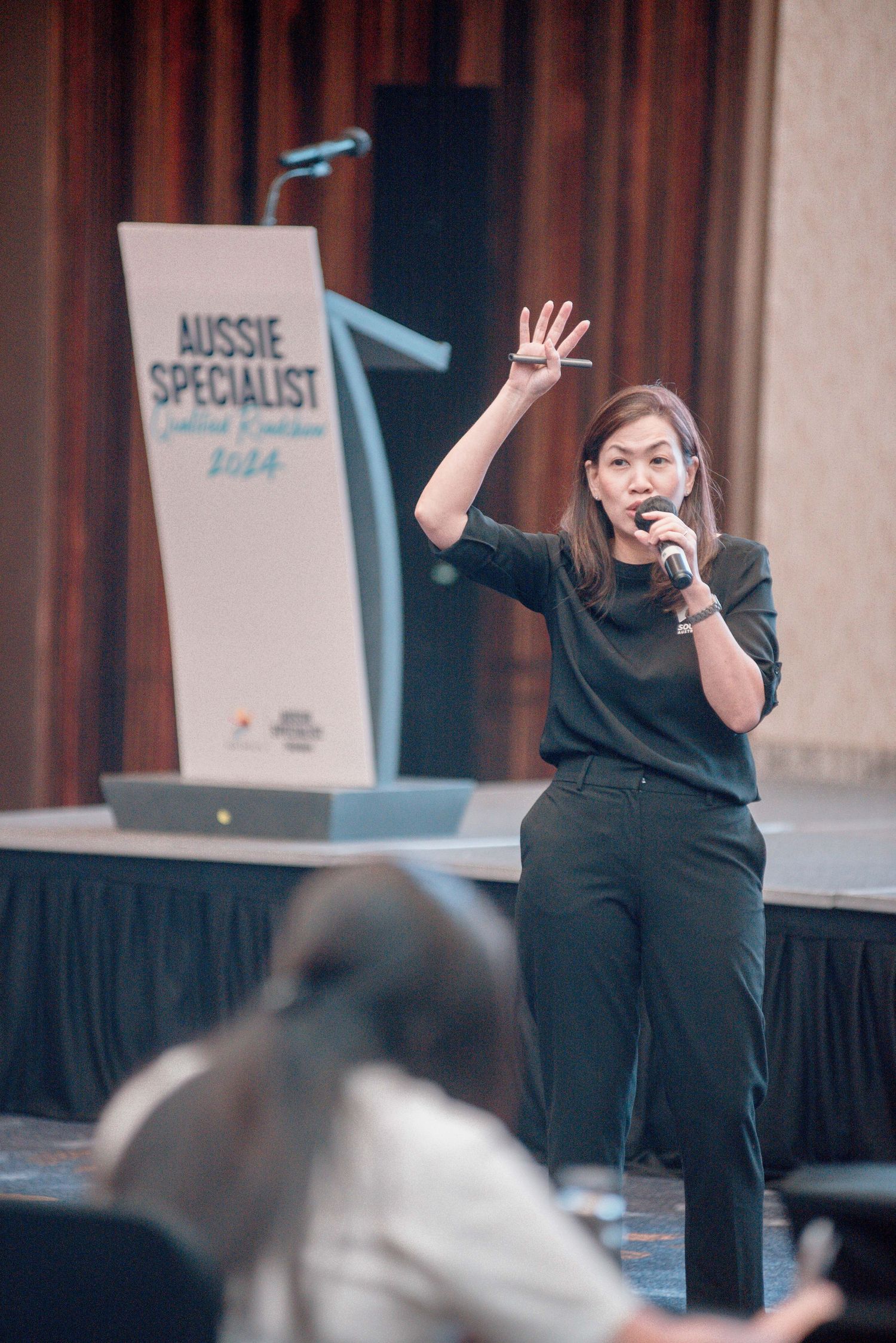 A speaker presents on stage at an Aussie Specialist event, wearing all black and gesturing enthusiastically to the audience.