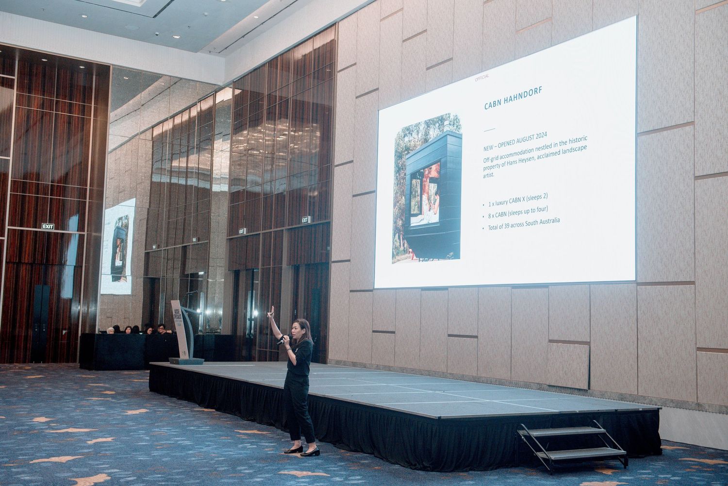 A presenter stands on a stage in front of a large projection screen in a modern conference room setting.