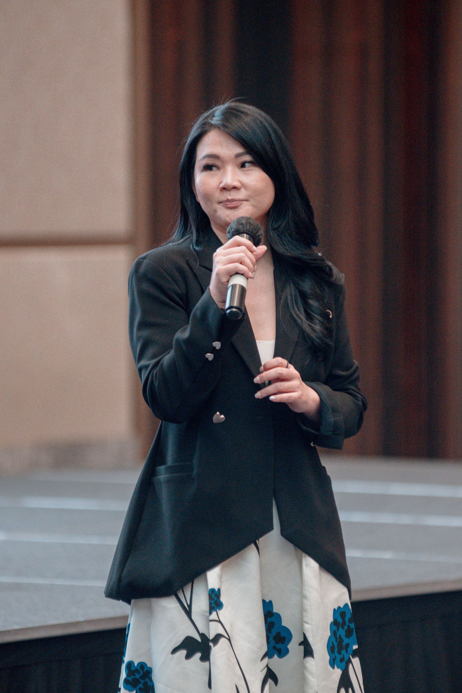 A woman in a black blazer and white dress with blue floral details stands outside a building.
