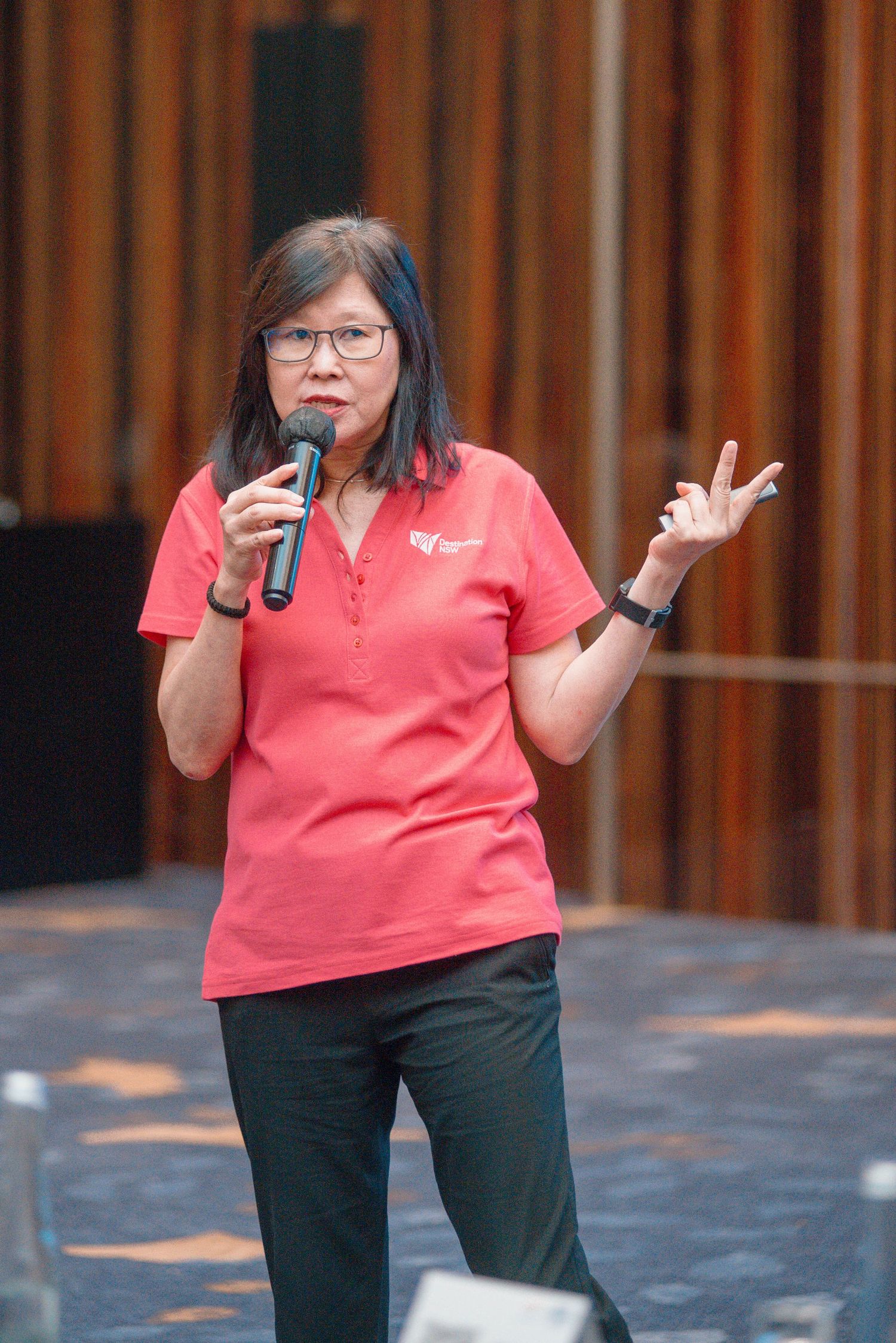 A person in a coral polo shirt and dark pants gestures while speaking on stage during a presentation.