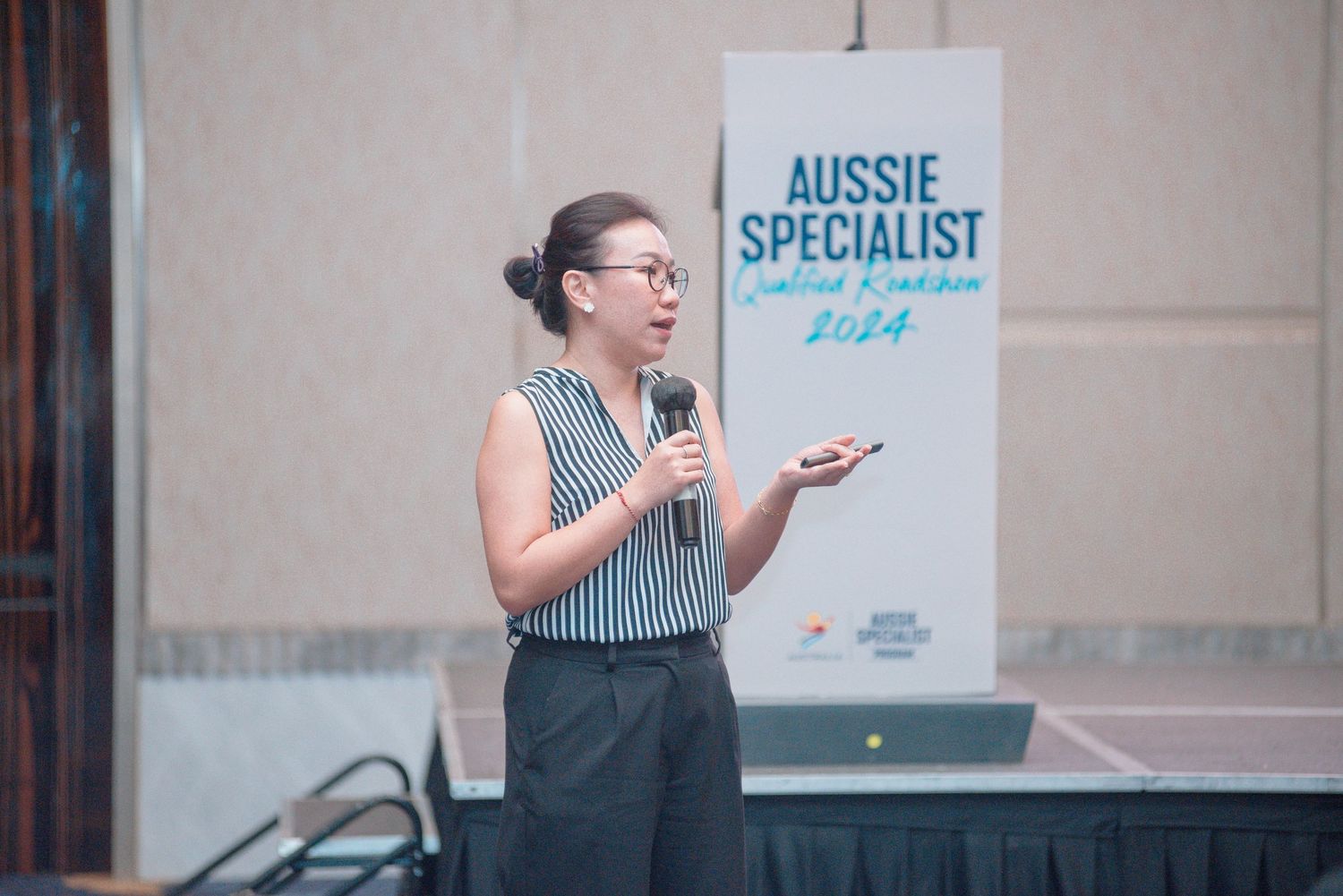 A person in a striped top gestures while speaking at a business presentation with signage visible in the background.