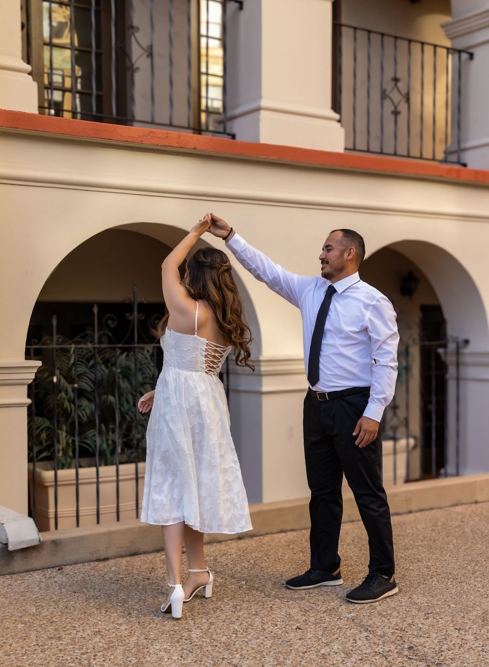 A couple performs a series of dance moves in a white dress and black suit against a beige building exterior.