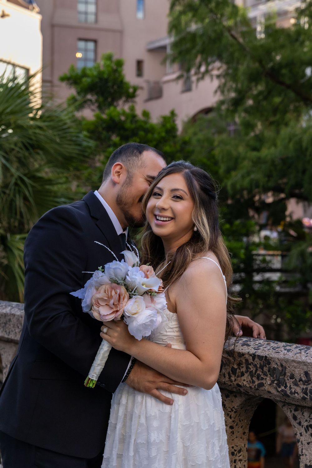 A couple shares an intimate moment outdoors, with the bride holding a soft pastel bouquet against a backdrop of palm trees.
