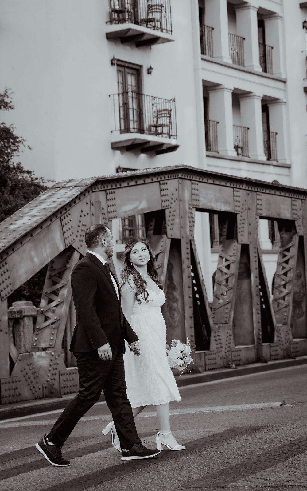 A black and white photo of a couple walking down a street with historic architecture and balconies in the background.