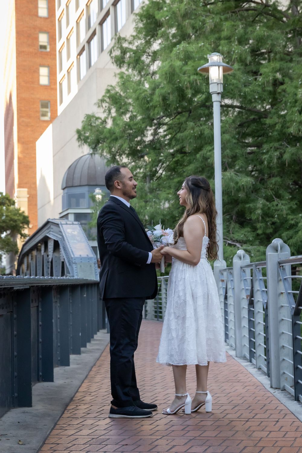 A couple in formal attire stands together on a brick walkway with railing and trees in an urban setting.