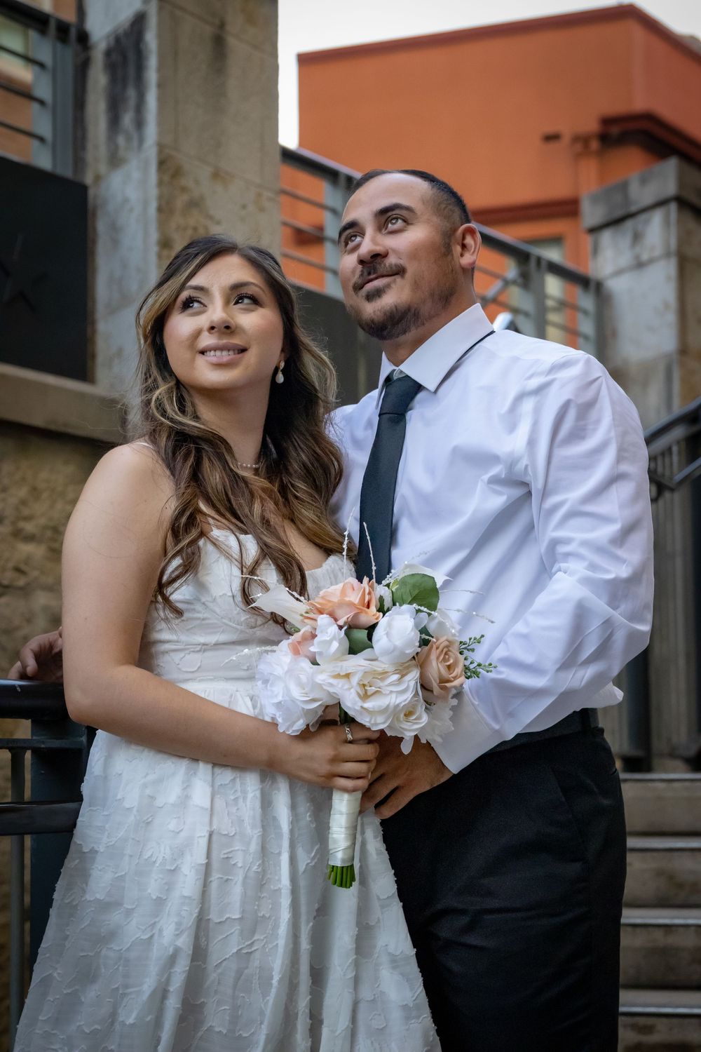 A couple in wedding attire smiles while standing on stone steps with the bride holding a light-colored bouquet.