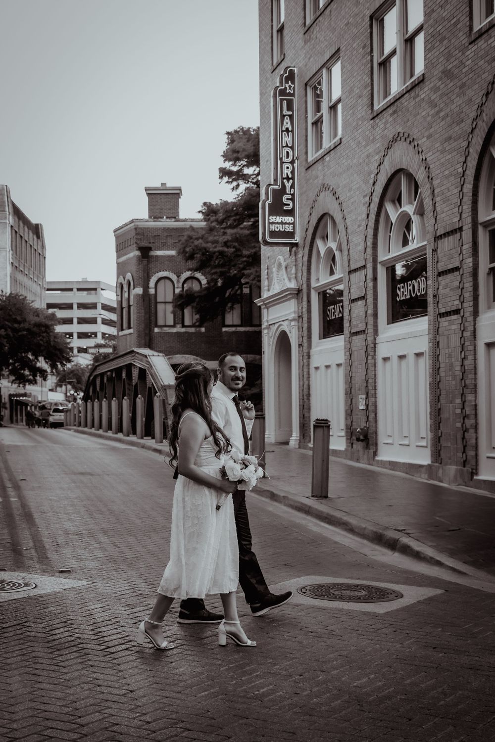 A couple stands on a brick street in a vintage-style black and white photograph near historic buildings.