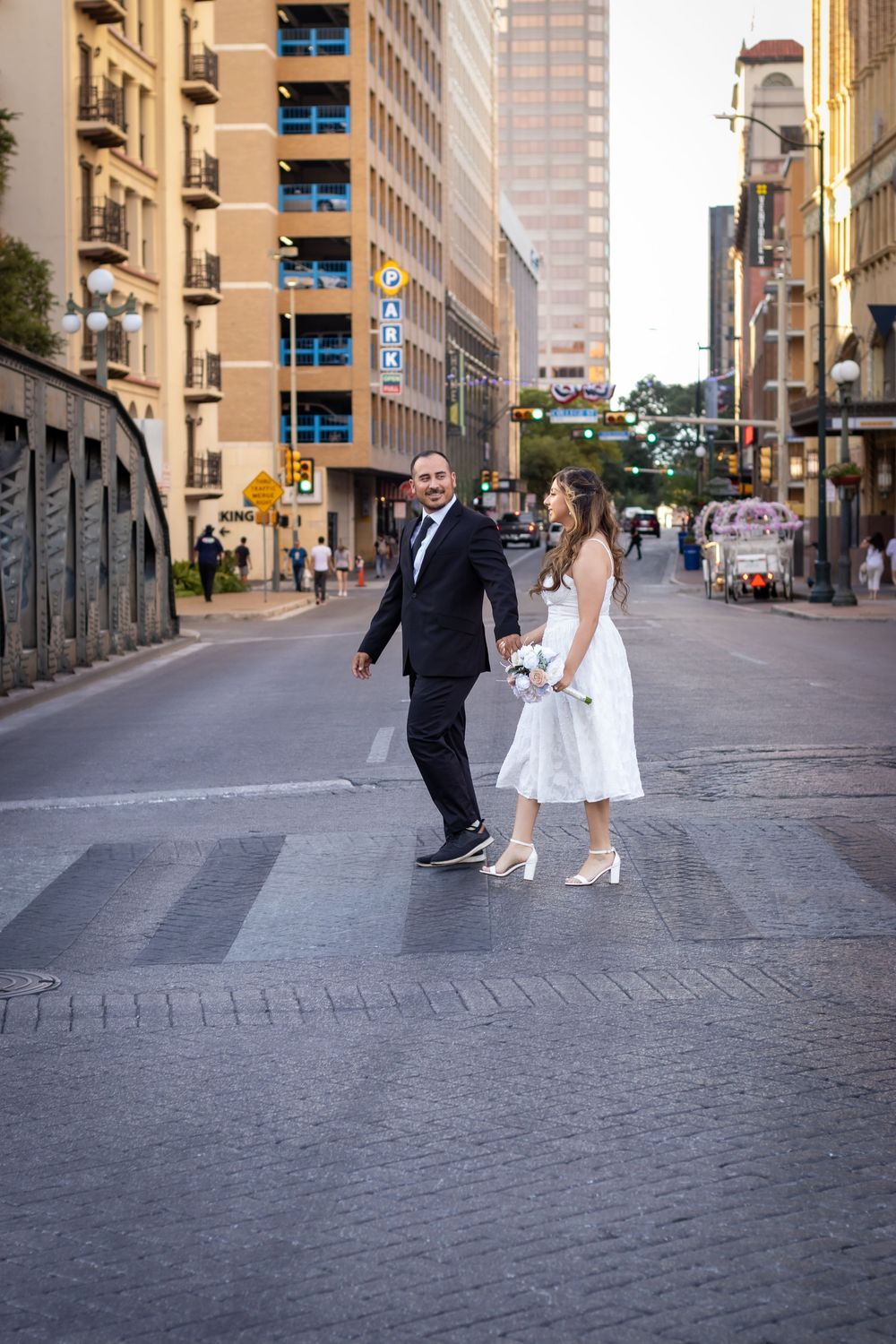 A couple in formal attire crosses an urban street in downtown Austin with tall buildings in the background.