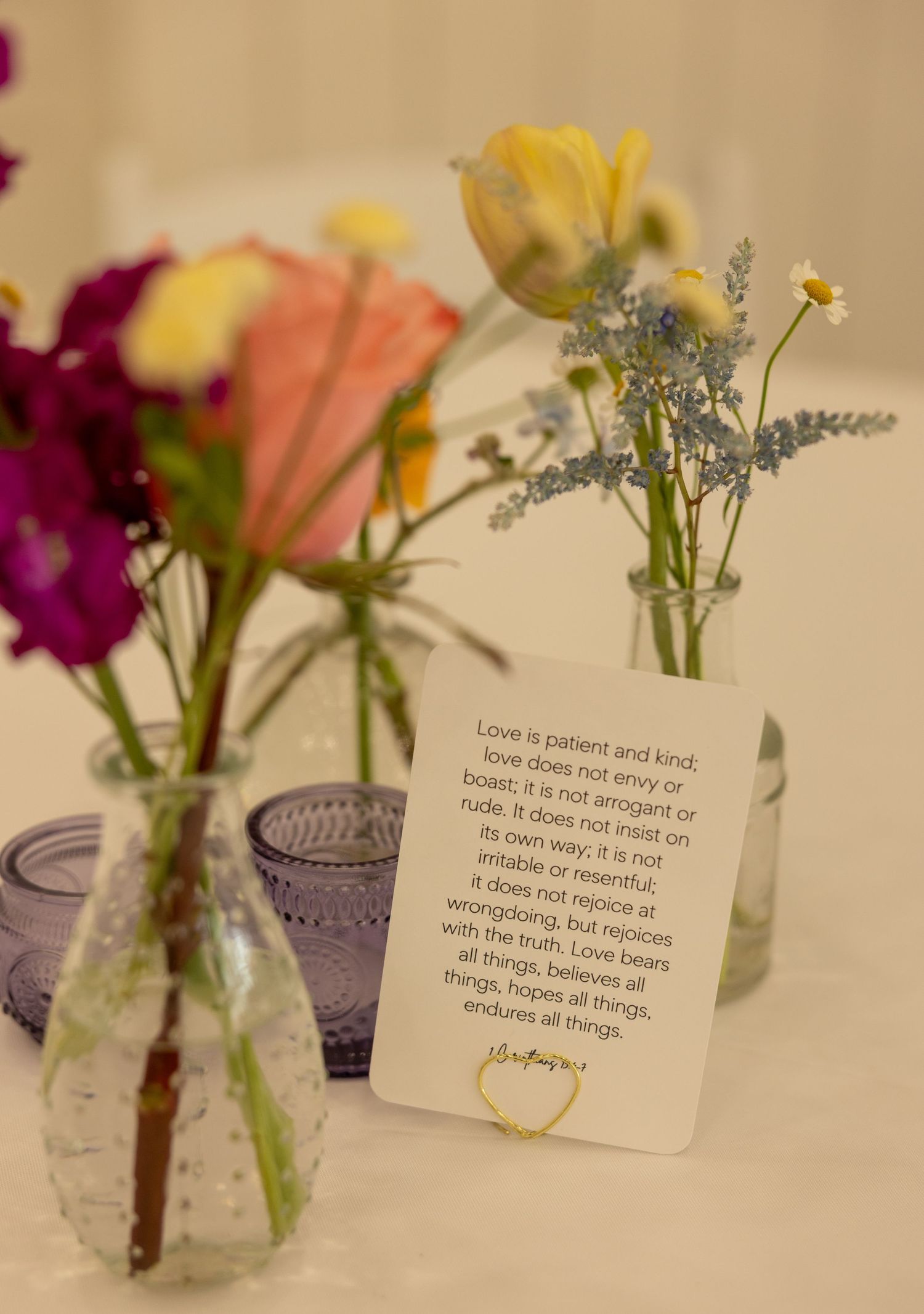 Wedding table display with colorful flowers in glass vases beside a 1 Corinthians 13 scripture card.