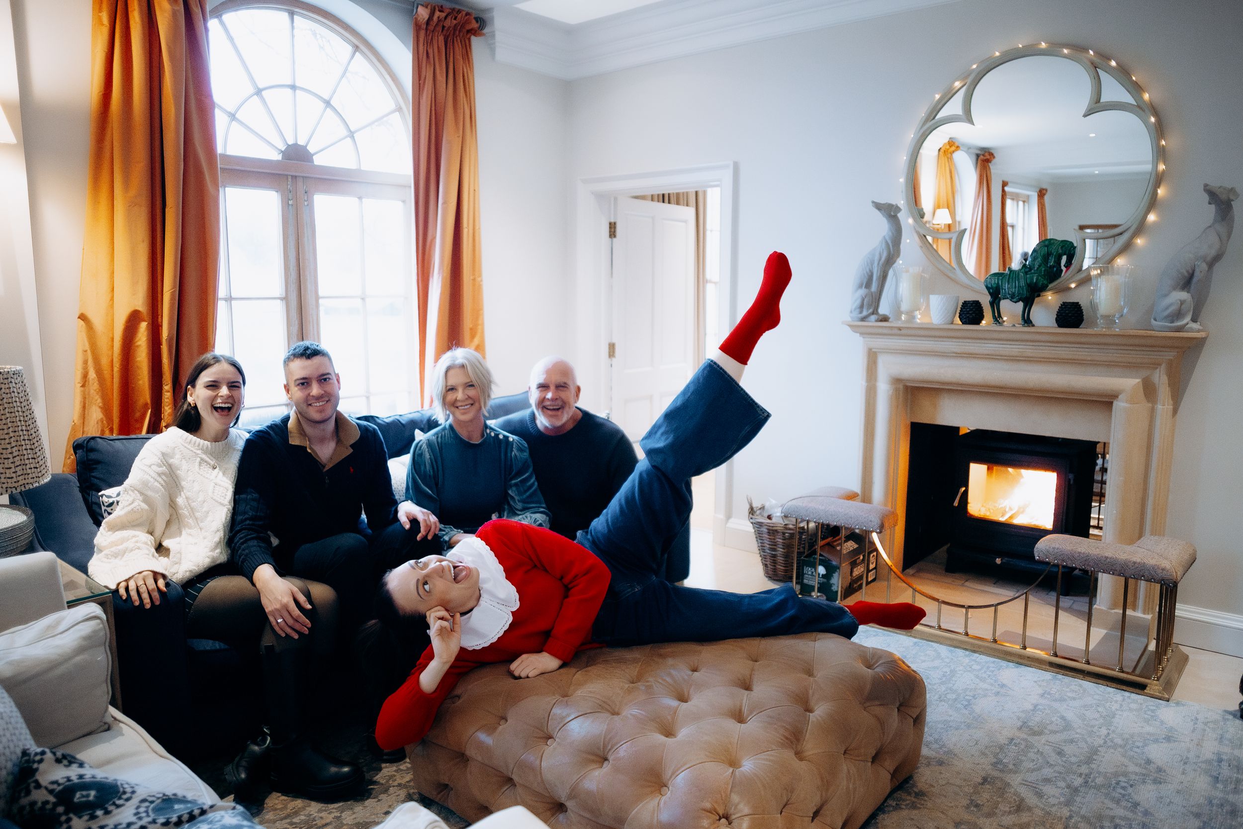A group of people lounging casually on a white couch in a bright living room with orange curtains.