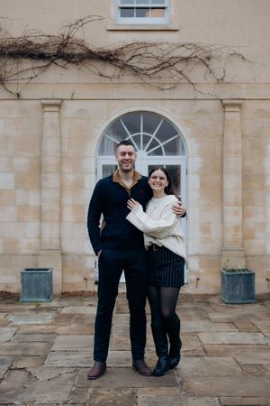A couple poses together against a stone wall with arched windows and climbing vines in a romantic architectural setting.
