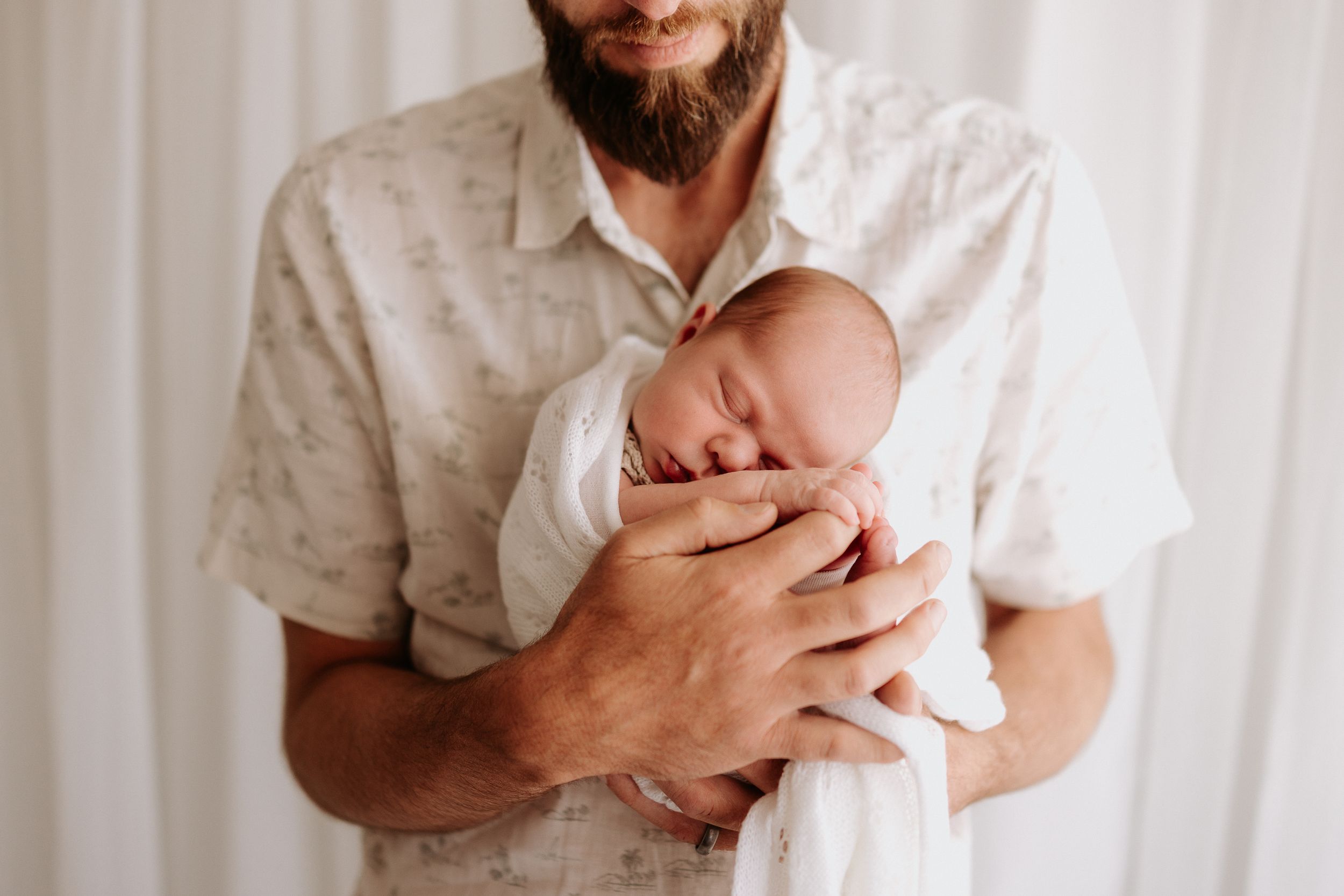 A sleeping newborn baby swaddled in white is gently cradled in protective arms against a white background.
