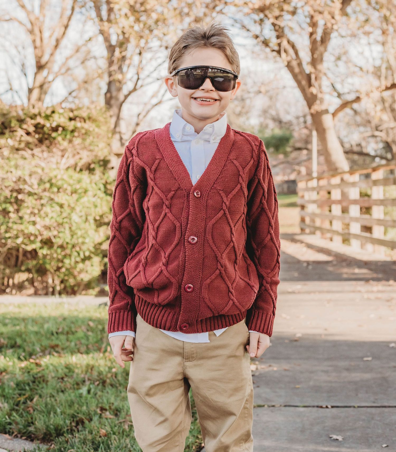 A young child wearing a maroon cable knit cardigan, khaki pants and sunglasses poses outdoors on a path.