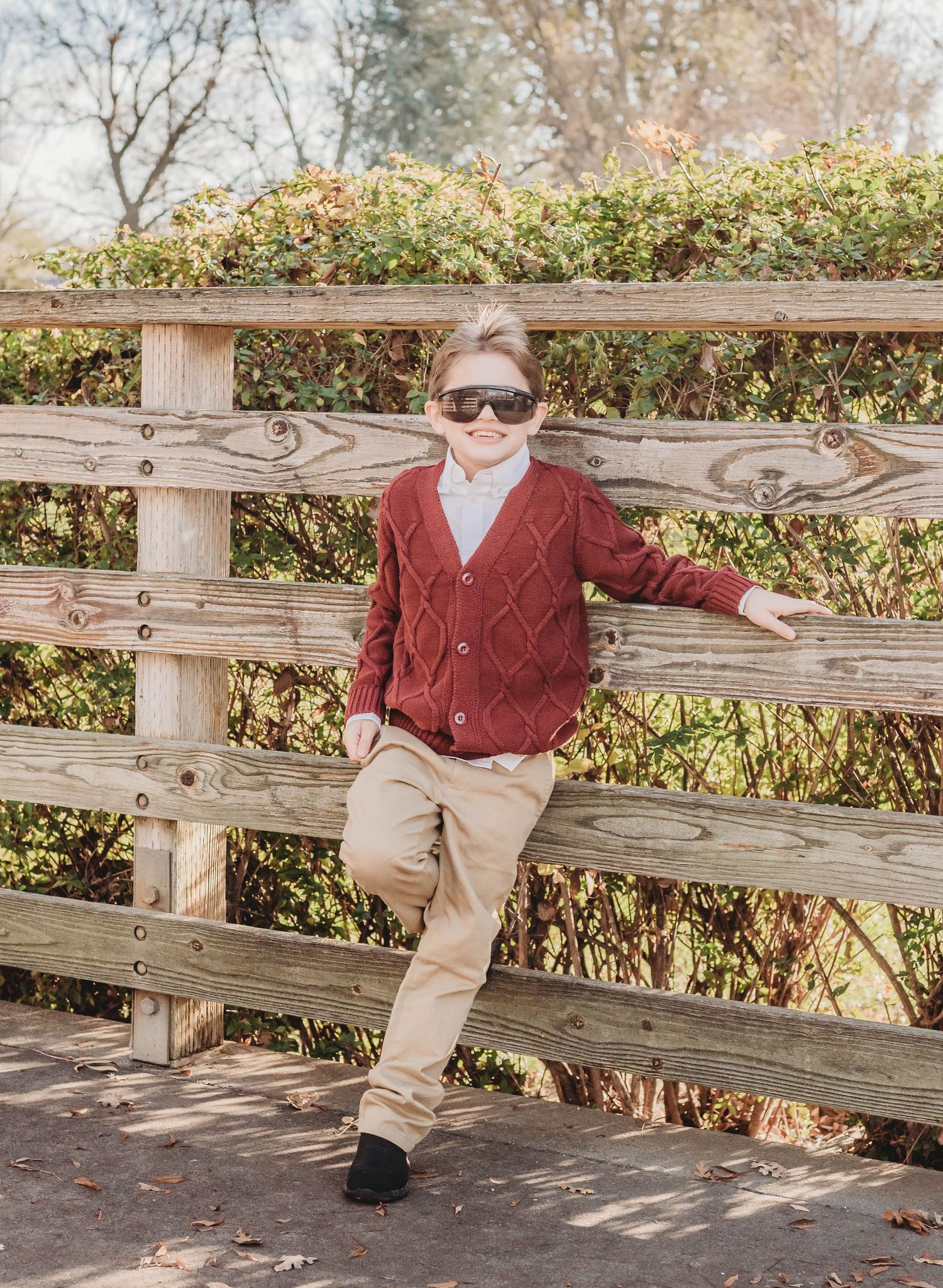 A young child in sunglasses and a red cardigan sweater leans casually against a wooden fence on a sunny autumn day.