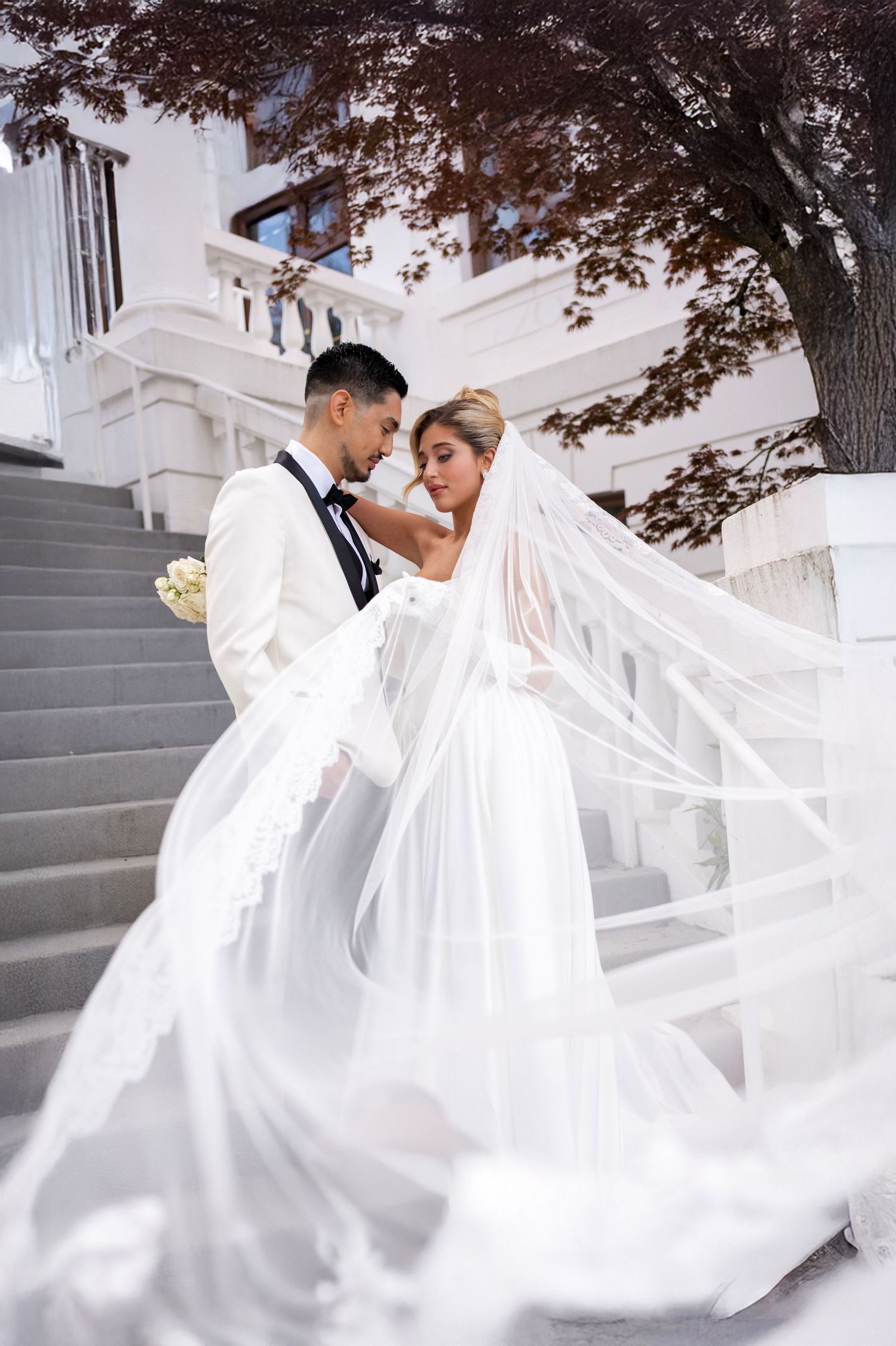 Bride and groom share a romantic moment on white stairs, her long veil flowing dramatically in the breeze.
