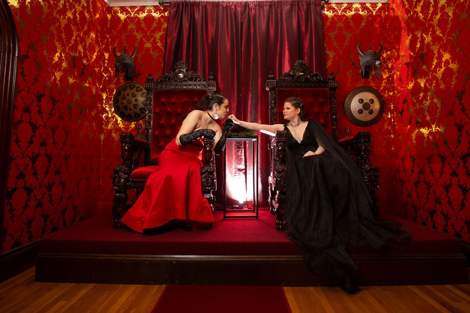A couple in formal attire sit in thrones in a luxurious red room with ornate wallpaper and dramatic lighting.