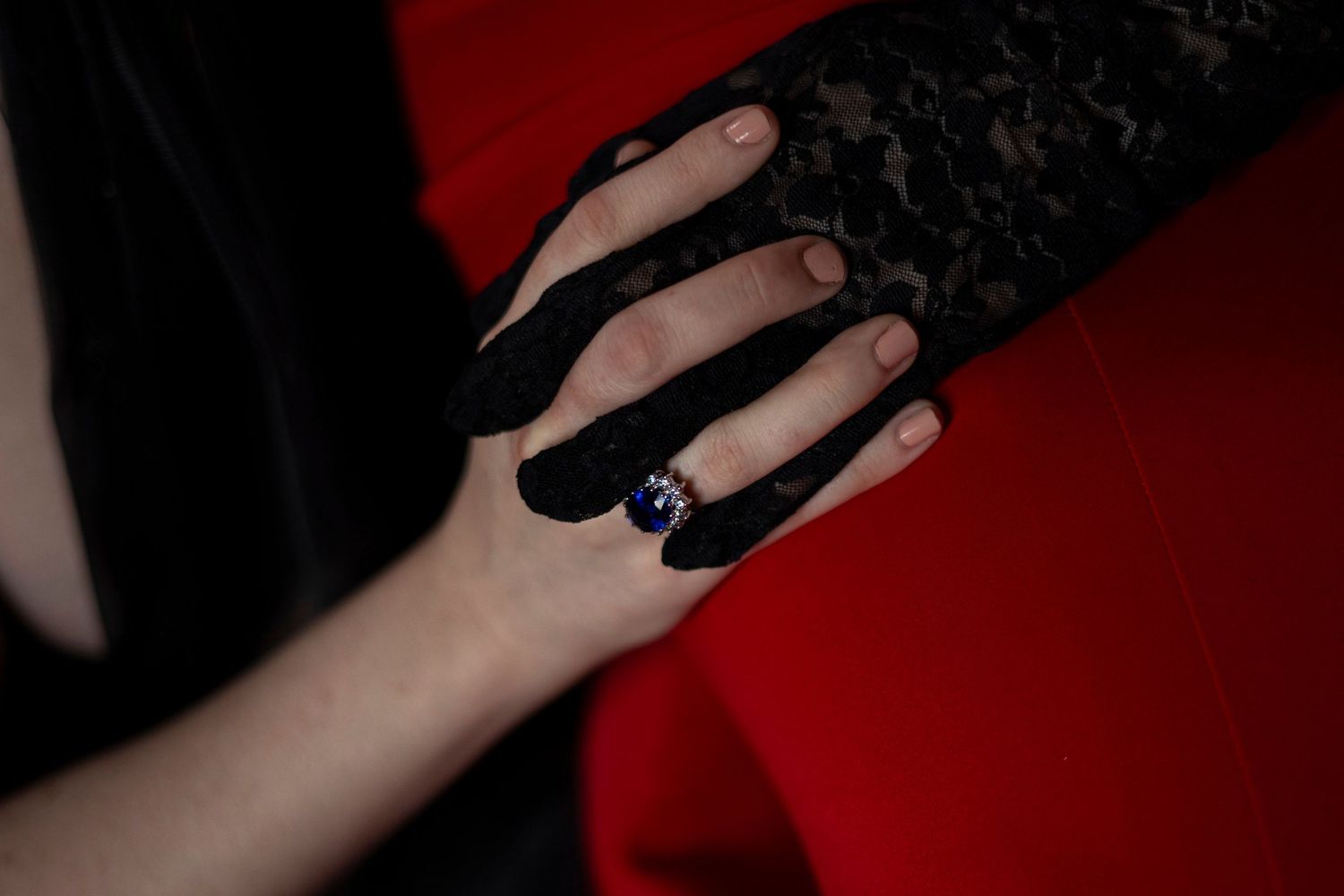 A hand wearing a black lace glove with a sapphire gemstone engagement ring holds the hand of her partner.