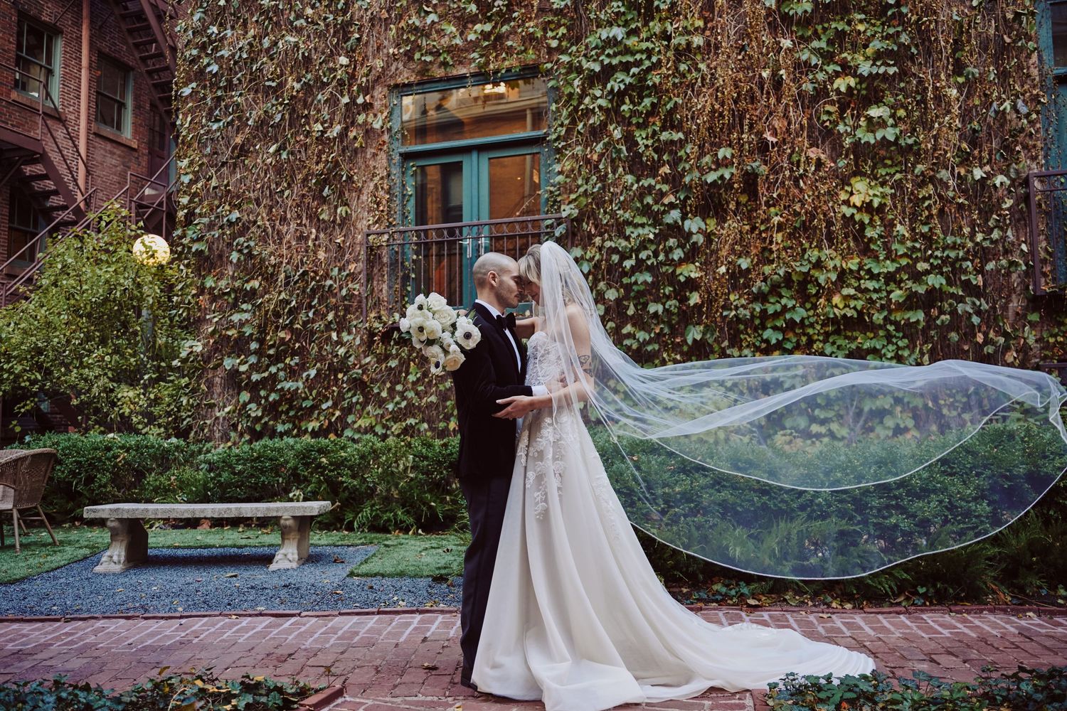 A couple in wedding attire embraces in front of an ivy-covered brick wall as a long veil flows dramatically in the breeze.