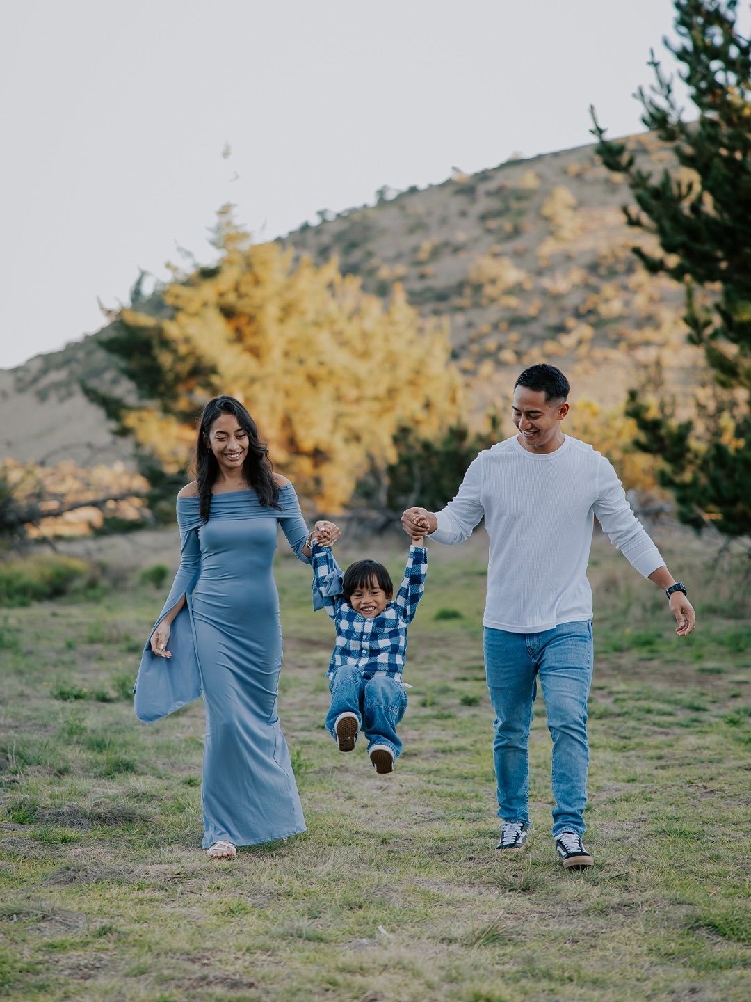 A family enjoys a playful moment together on a grassy hillside during golden hour with autumn trees in the background.