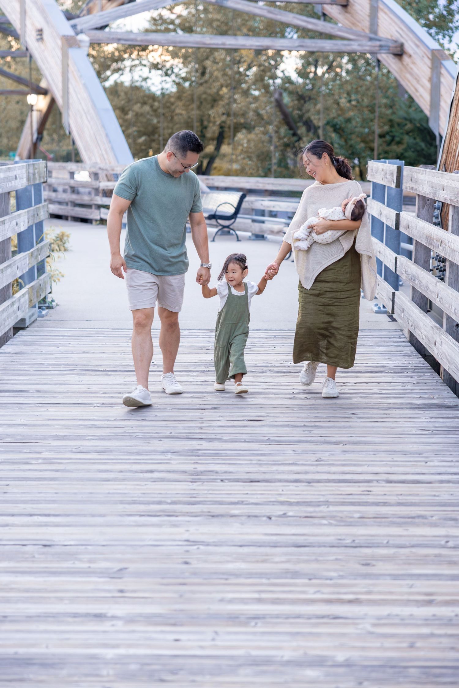 Family walking together on a wooden boardwalk bridge surrounded by trees on a sunny day.